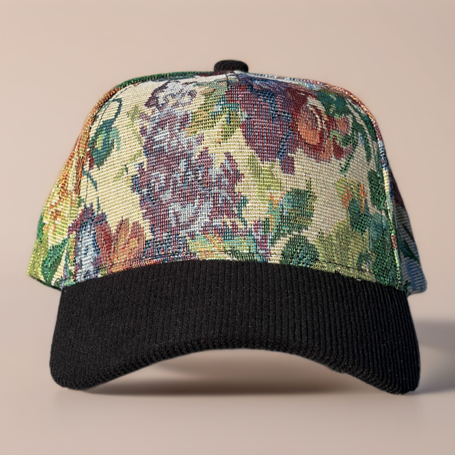 A baseball cap with a black ribbed brim and a front panel featuring a dense floral tapestry pattern in green, red, blue, and yellow tones, set against a solid beige background.