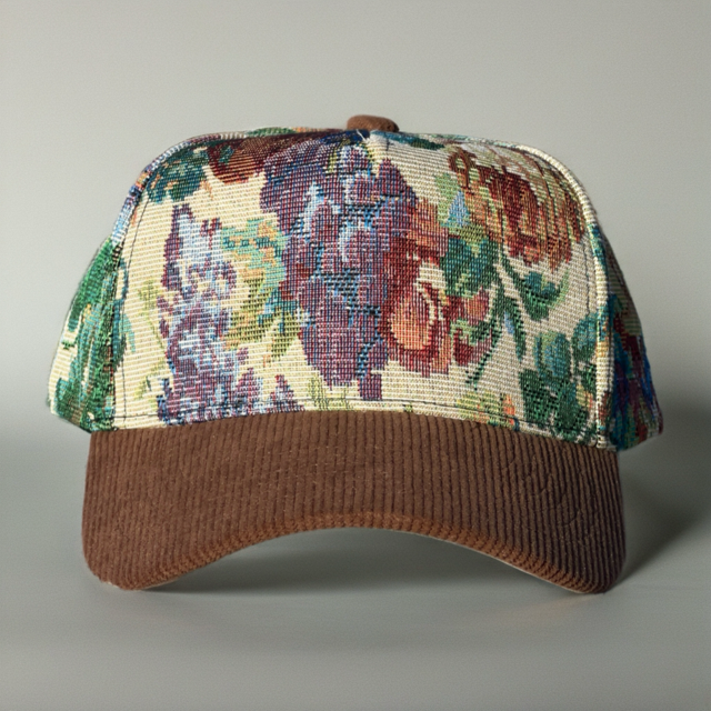 A baseball cap with a brown corduroy visor and a front panel featuring a dense floral tapestry pattern in purple, green, red, and cream, photographed against a neutral gray background.