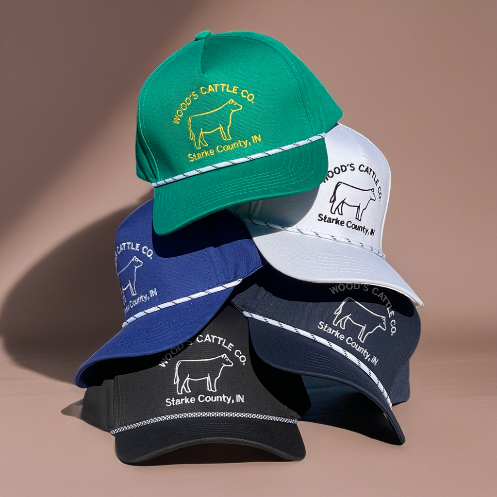 A stack of six Wood's Cattle Co. baseball caps in various colors (green, white, blue, navy, black) with embroidered logos and cow illustrations, set against a solid beige background.