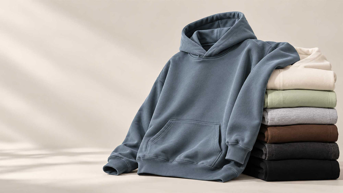 A slate blue pullover hoodie is draped over a stack of folded sweatshirts in cream, sage green, heather gray, brown, and black, set against a warm beige background with soft diagonal shadows.