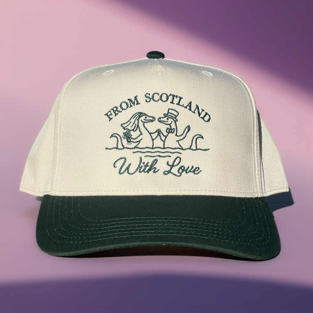 A trucker-style baseball cap with an off-white crown and a dark green brim, featuring embroidered text and a dachshund illustration on the front. The text reads 'FROM SCOTLAND With Love'.