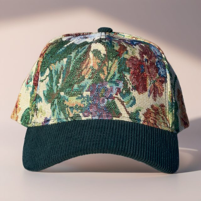 A baseball cap with a dark green ribbed brim and a front panel featuring a detailed floral tapestry pattern in green, red, blue, and beige, set against a warm beige background with soft shadows.